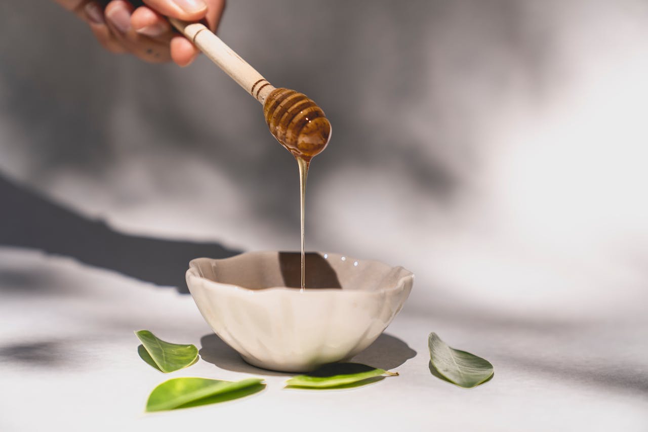 Close-up of honey flowing from a dipper into a ceramic bowl, surrounded by green leaves.