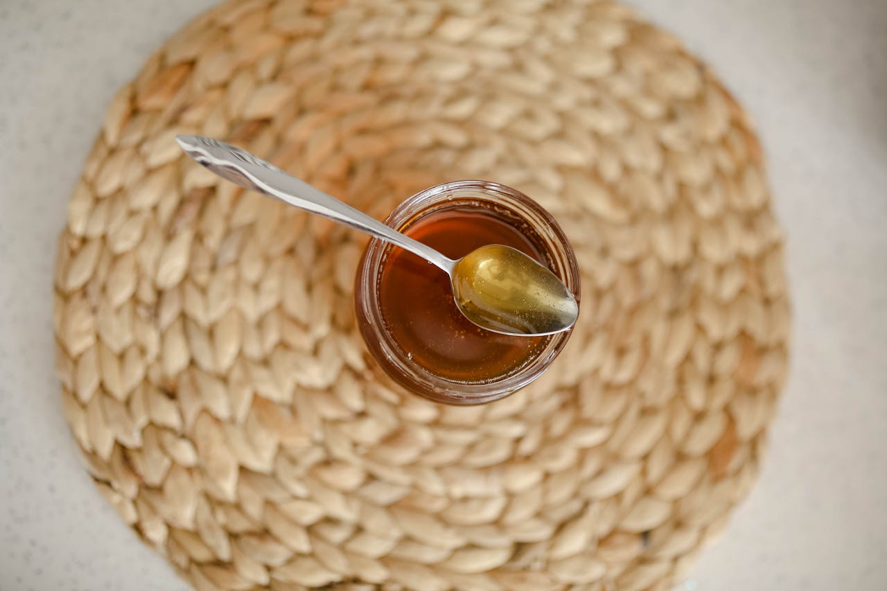 Overhead view of a honey jar with a silver spoon on a braided placemat, showcasing simple elegance.